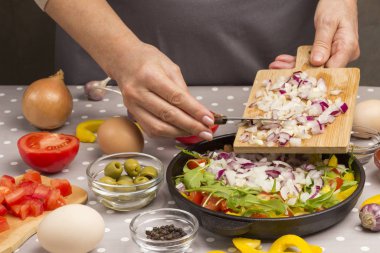 Tavada dilimlenmiş sebze. Eller tavaya soğan koyar. Zeytin, yumurta.,