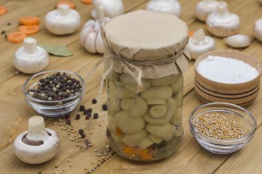 Fermentation Products. Glass jar with canned mushrooms and fresh champignon mushrooms. Spice: salt, garlic, onion, bay leaf on table. Healthy winter foods, Copy space.  Light wooden background