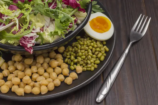 Multicolored leafy vegetables mix, chickpeas, mung bean, egg and fork in black plate. Balanced nutrition. Dark wooden background