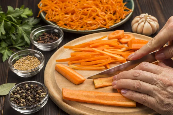 Hands chop raw carrots on cutting board. Spices, garlic and parsley on table. Fermented carrots in plate. Natural cold remedy. Close up. Dark wooden background.