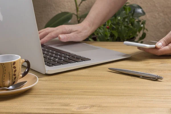 Hand is typing on laptop, second hand is holding smartphone, pen and cup of coffee on light wooden table. Work from home