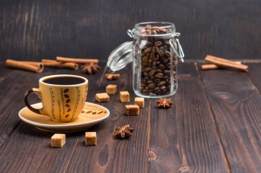 Bir fincan kahve, cam kavanozlarda kahve çekirdekleri. Tarçın çubuklarında, yıldız anasonunda ve esmer şeker parçalarında. Koyu tahta arkaplan