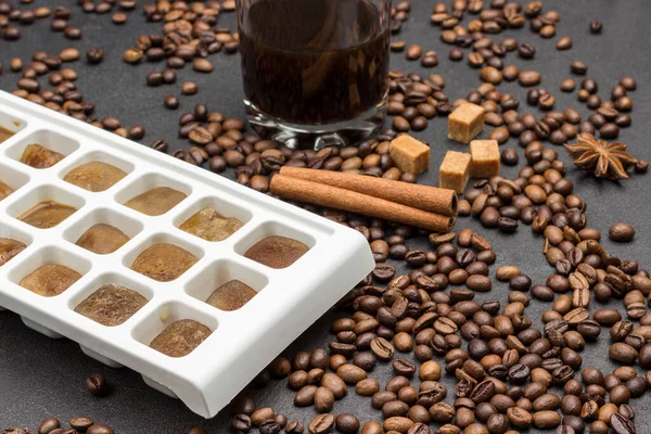 Coffee ice cubes. Glass with coffee. Coffee beans scattered on table, star anise, cinnamon sticks and pieces of brown sugar. Dark background