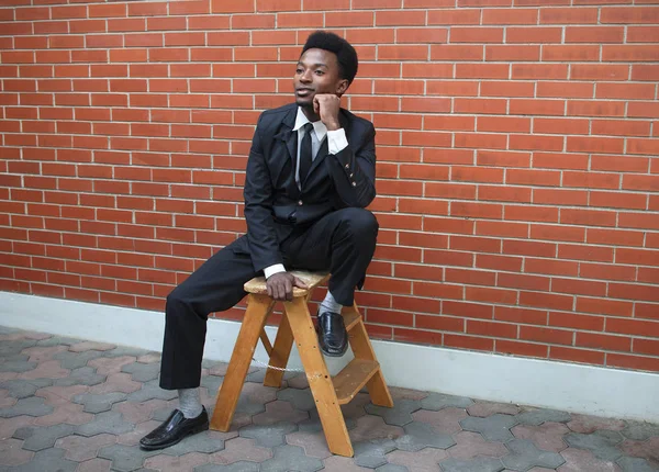 Fiatal férfi öltöny és a nyakkendő ül téglafal — Stock Fotó