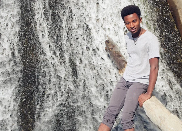 Unga afrikanska man sittande på trädstam i floden vattenfall — Stockfoto