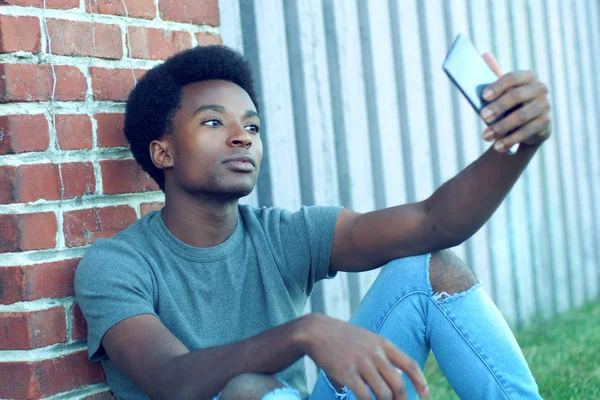 Ung man tar selfie utanför sitter i gräset bär jeans smartphone pic porträtt teknik — Stockfoto