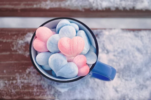 pink and blue meringue hearts in blue metal mug on a wooden brown bench with snow, winter