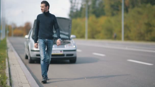 Un hombre sentado en la carretera delante de un coche roto — Vídeo de stock