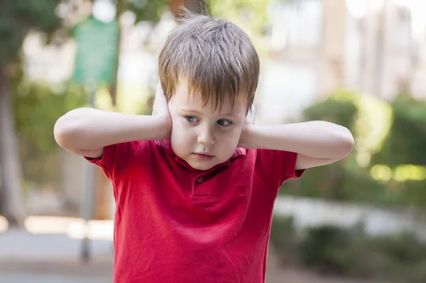 Pequeno menino caucasiano em camisa de pólo vermelho fechando as orelhas com as mãos e olhando para o lado . — Fotografia de Stock