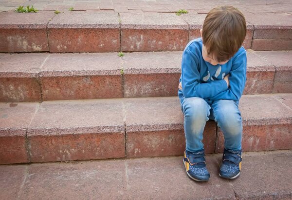 Unhappy little Caucasian child sitting on the stairs in a closed position with his head down.