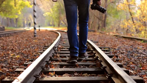 Um homem com câmera DSLR desce trilhos de trem em uma floresta de outono de fundo. Um homem sai da câmara. — Vídeo de Stock