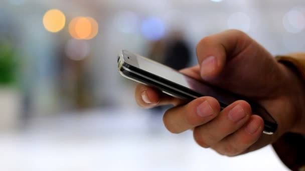 Una mano de hombre utiliza un teléfono inteligente negro en el fondo caminando personas y luces bokeh — Vídeo de stock