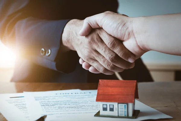 Estate agent shaking hands with his customer after contract signature, Contract document and house model on wooden desk
