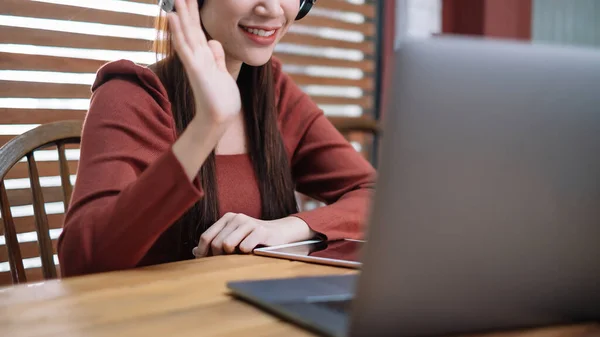 Smiling Asian businesswoman in headset waving hand, using laptop, looking at screen, student learning online