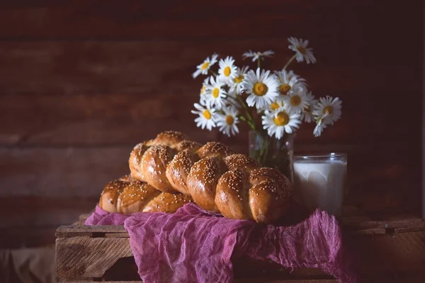 Newly-baked wheat long loaf on wooden stoleevreysky challah. Long loaf a braid with sesame on a table with a glass of milk and a bouquet of camomiles. A still life with bread and flowers