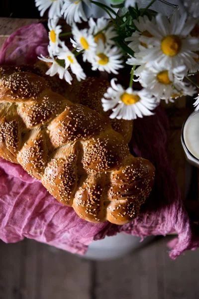 Newly-baked wheat long loaf on wooden stoleevreysky challah. Long loaf a braid with sesame on a table with a glass of milk and a bouquet of camomiles. A still life with bread and flowers