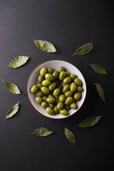 Green olives in a white bowl on a black background. Background of olives. Background with green olives. Olives. Bay leaf.