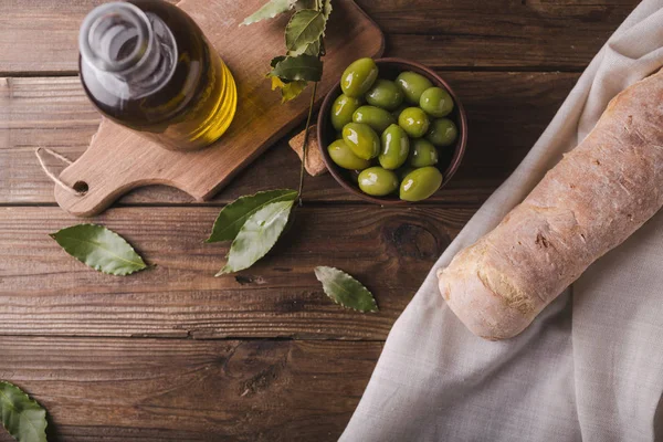 Aceitunas verdes sobre una tabla de madera ciabatta, aceite de oliva, hoja de laurel sobre fondo de madera. Ciabatta, copyspace — Foto de Stock