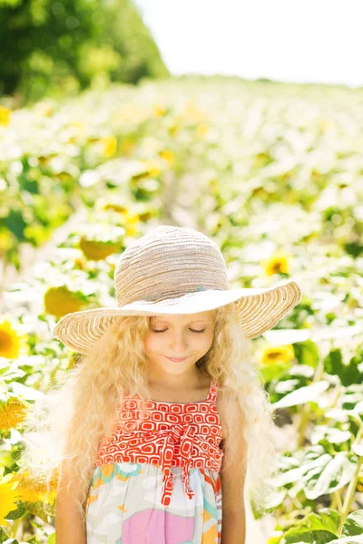 Ayçiçeği tarlası kıvırcık beyaz saçlı küçük kız. Kız bir hasır şapka ve sarafan