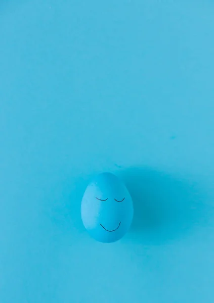 Blue Easter egg funny painted face on blue background. Copy space.