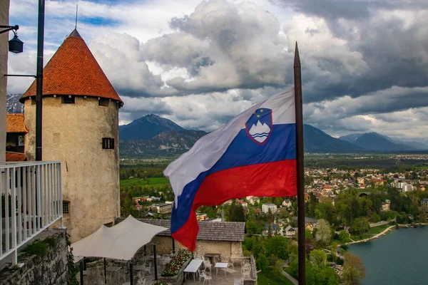 Slovenian national official flag waving on flagpole of Bled Castle, Slovenia Tower of the castle covered red tiling. Top view on famous Bled town and Karavanke mountain range on background