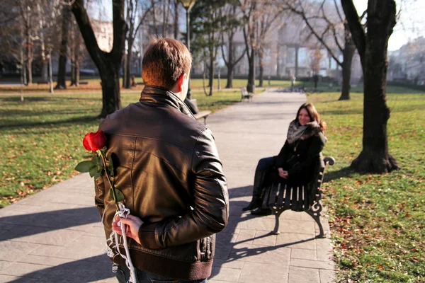 Mädchen sitzt im Park und Freund nähert sich mit roter Rose — Stockfoto