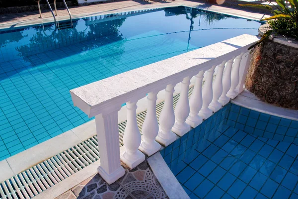 Luxury pool with white balusters. Empty swimming pool with classic balustrade, shower and chaise lounge. Summertime, holiday, travel, water with sunny reflections. Architectural decorative elements.