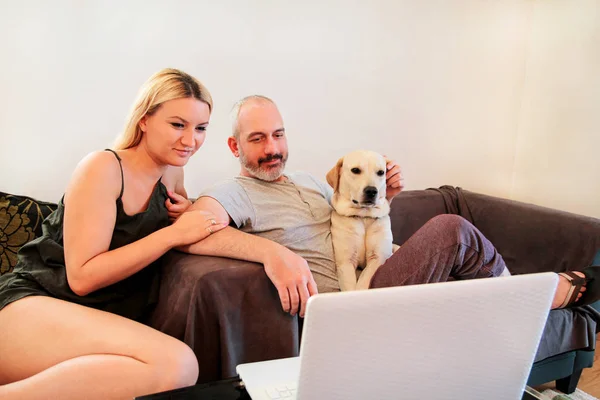 Lidé se psem doma. Mladý šťastný pár si se svým psem žlutý labrador retrívr na dům obývací pokoj, pomocí přenosného počítače v gauči. Hezký kluk a holka vlastníci mazlit a kterým se pejsek. — Stock fotografie