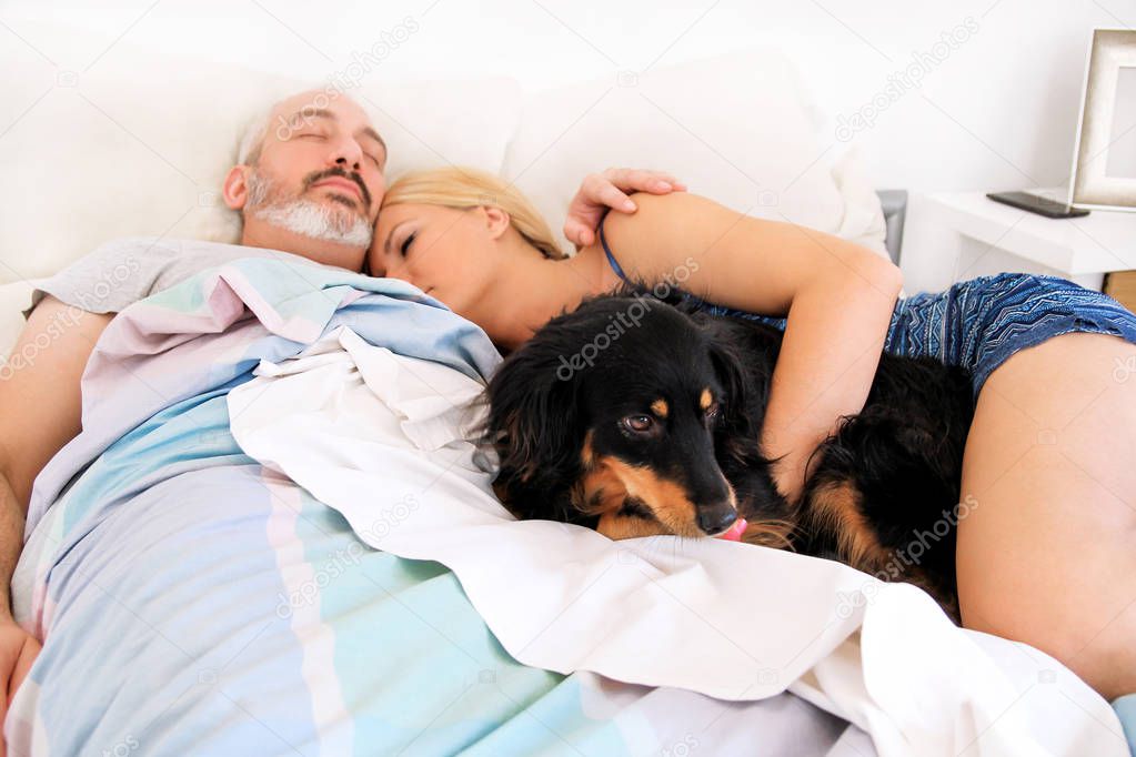 Handsome couple in bed sleeps together in association with their dog. A mixed breed dogs enjoys and resting in the bedroom with their owners. People with pets at home, house. Lovely dog, pet concept.