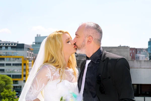 Concepto de boda. La pareja de novios guapos felices están de pie y besándose en el techo del edificio. Los novios enamorados se abrazan. En el fondo están los edificios, escena urbana de la ciudad . —  Fotos de Stock