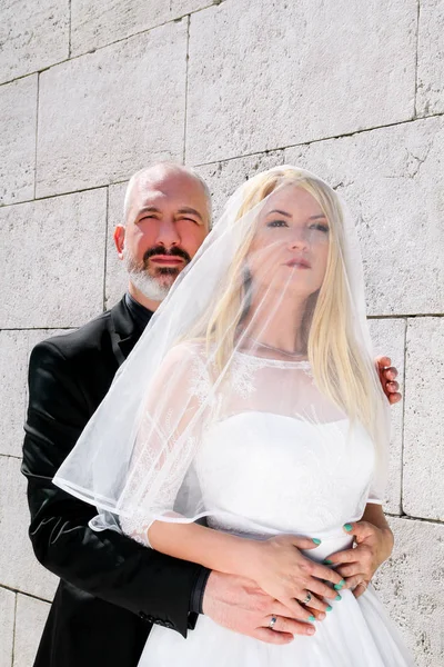 Concepto de boda. Una pareja de casados guapos se encuentra junto a la pared de piedra blanca, gente encantadora mirando a la distancia y posando hacia la cámara. El hermoso novio y la novia en el amor abrazo . —  Fotos de Stock