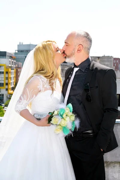 Concepto de boda. La pareja de novios guapos felices están de pie y besándose en el techo del edificio. Los novios enamorados se abrazan. En el fondo están los edificios, escena urbana de la ciudad . —  Fotos de Stock