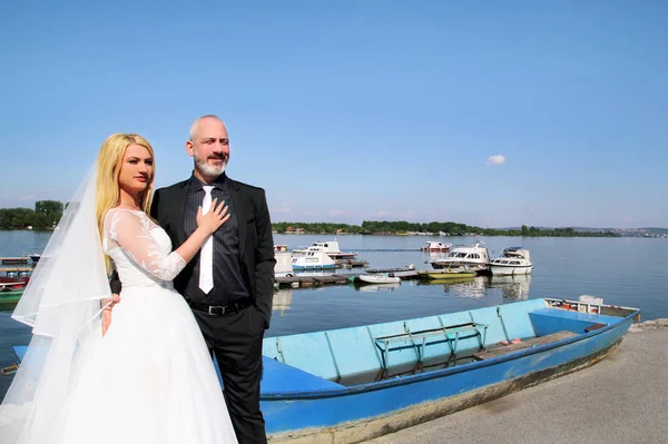 Wedding concept. The happy handsome wedding couple are sitting and looking. The groom and bride in love embrace. In background is beautiful natural environment, a river with fishing boats. Happiness.