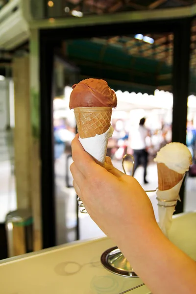 The female hand holds ice cream in the corner. Ice cream in waffle cone. Fridge with ice cream in the background. Woman works in ice cream shop. Pastry shop. Vanilla, chocolate, fruit. Shop of sweets.