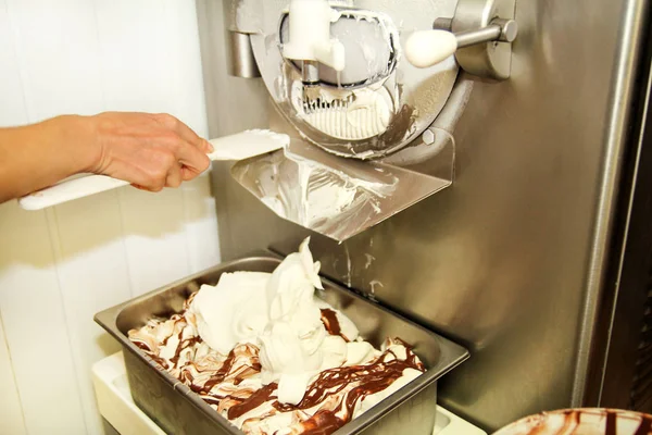 Female worker is working on ice cream maker machine. Producing vanilla ice cream with chocolate dressing and it falls into steel container. Industrial preparation of creamy ice cream. Dessert, sweets.
