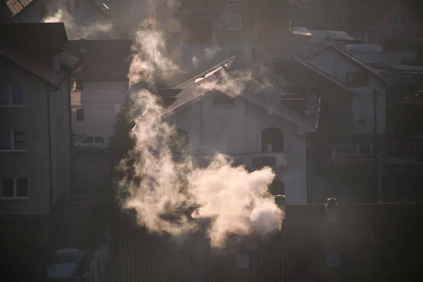 Smoking chimneys at roofs of houses emits smoke, smog at sunrise, pollutants enter atmosphere. Environmental disaster. Harmful emissions and exhaust gases into air. Fog, winter day, heating season. Stock Photo