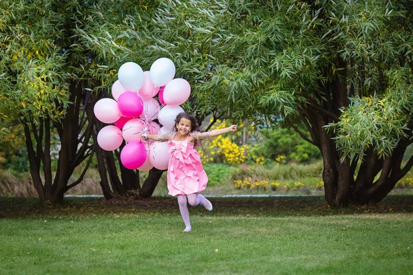 Menina encantadora em um vestido rosa inteligente corre para se encontrar com um grande grupo de balões rosa . — Fotografia de Stock
