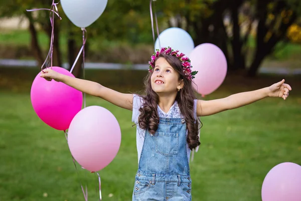 Hermosa niña de pelo oscuro en overoles de mezclilla de pie en el césped rodeado de globos rosados y blancos — Foto de Stock