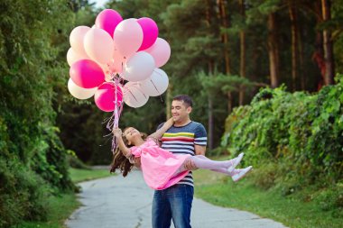 Baba küçük kızı ellerinde tutar. Bir kız güzel pembe elbise giymiş ve renkli balonlar büyük bir sürü holding 