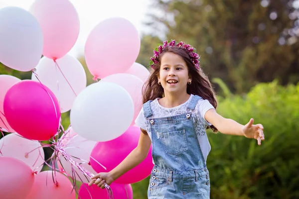 Смеющаяся красивая девушка в джинсовом комбинезоне и цветочный венок бегут на встречу с большой связкой розовых шариков. Портрет, избирательный фокус . — стоковое фото