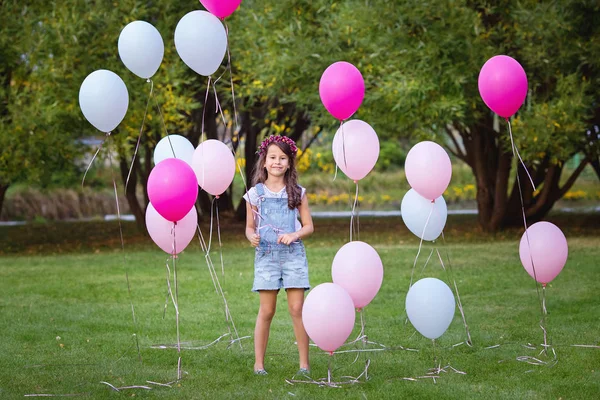 Красивая темноволосая маленькая девочка в джинсовом комбинезоне и с цветочным венком на голове стоит на лужайке в окружении розовых и белых шариков — стоковое фото
