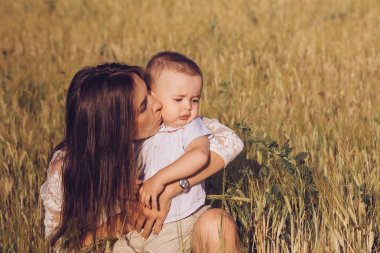 Sevimli genç esmer anne hugs ve küçük oğlu bir yaz alanında oturup kisses