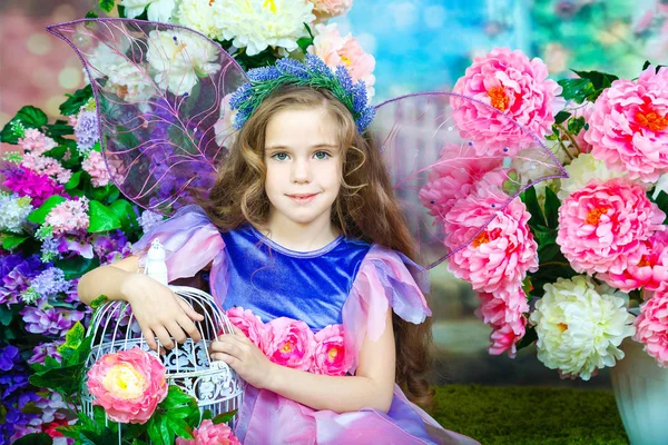 Ritratto di una graziosa bambina con i capelli ricci in un abito fata colorato con ali trasparenti, seduta tra i fiori accanto a una gabbia decorativa per uccelli in pizzo. Studio fotografia — Foto Stock