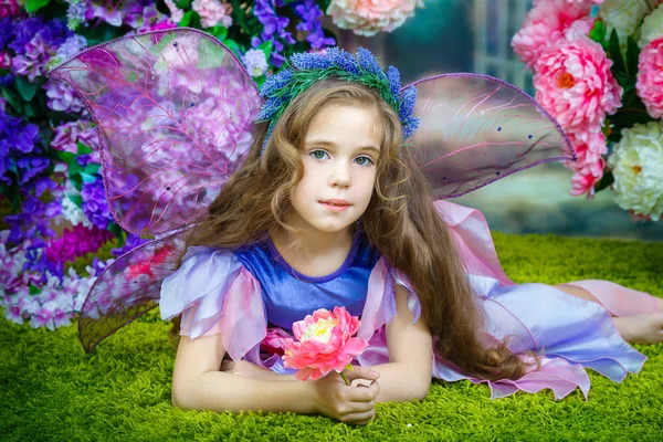 Портрет очаровательной маленькой девочки с вьющимися волосами в красочном сказочном платье с прозрачными крыльями, лежащими на травянистом зеленом ковре. Студийные фотографии . — стоковое фото