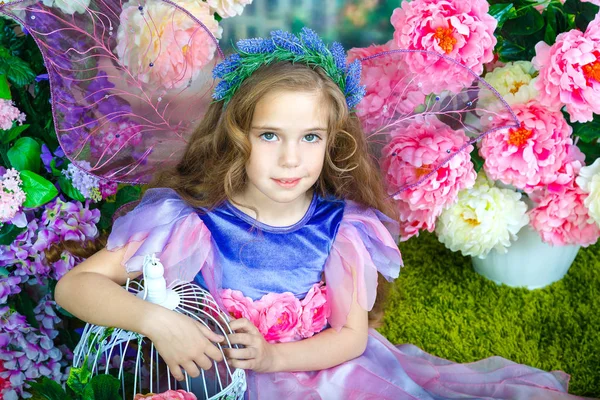 Портрет красивой маленькой девочки с вьющимися волосами в красочном сказочном платье с прозрачными крыльями, сидящими среди цветов на зеленом травяном ковре. Студийные фотографии — стоковое фото