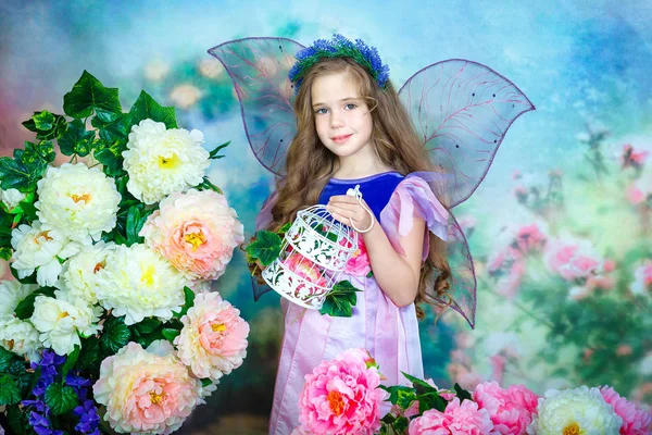 Retrato de una encantadora niña con el pelo rizado en un colorido vestido de hadas con alas transparentes se encuentra entre las flores de colores y sostiene una jaula de encaje decorativo en sus manos. Estudio fotografía . — Foto de Stock