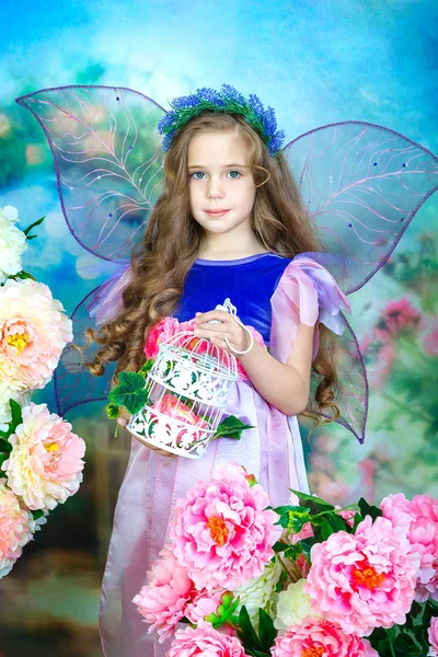 Portrait of a charming little girl with curly hair in a colorful fairy dress with transparent wings. The girl stands among the colorful flowers and holds a decorative lace birdcage in her hands. Studio photography. — Stock Photo, Image