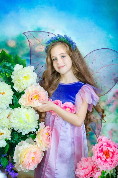 Ritratto di una graziosa bambina con i capelli ricci in un abito colorato fata con ali trasparenti. Una piccola fata si erge tra i grandi fiori . — Foto Stock