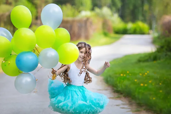 Pequeña chica sonriente hermosa con el pelo rizado largo usando falda mullida inteligente jugando en el jardín de primavera sosteniendo en las manos un gran montón de globos. Concepto de infancia feliz — Foto de Stock