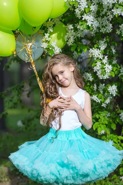 Pequena menina sorridente bonita com cabelos longos encaracolados vestindo saia fofa inteligente jogando no jardim da primavera segurando nas mãos um grande monte de balões. Conceito de infância feliz — Fotografia de Stock